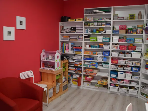 Sala terapeutyczna gabinetu: czerwona ściana, regały z pomocami logopedycznymi i fotel